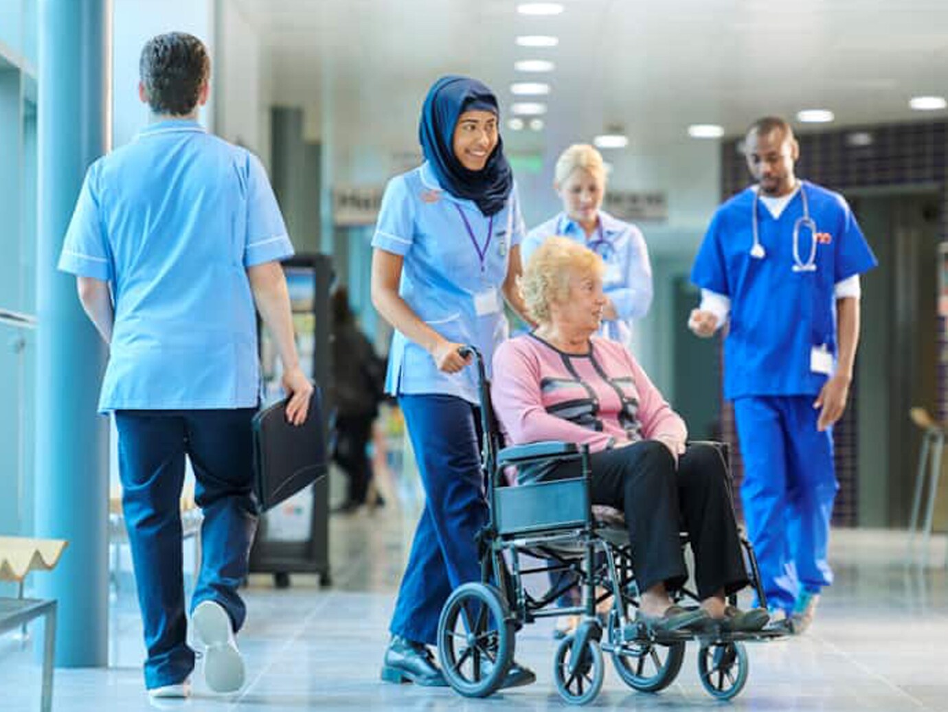 Nurse pushing an elderly man's wheelchair, while doctors and nurses walk in the background