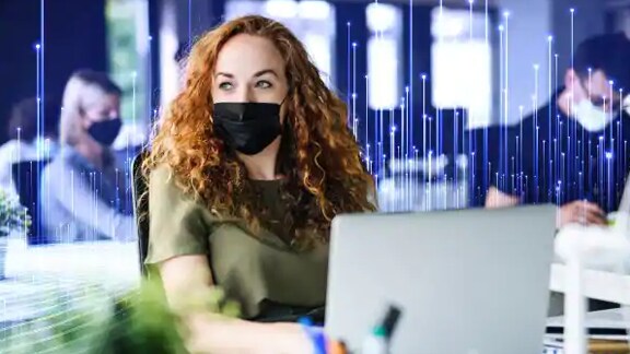 A woman wearing a facemask with a laptop at her desk, with people working on laptops in the background