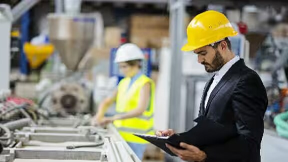 supervisor in safety helmet examining a document as a woman works at a distance in a factory