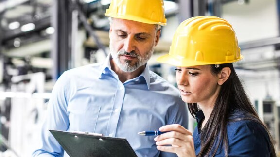 man and woman in safety helmets discussing work while looking at a writing pad