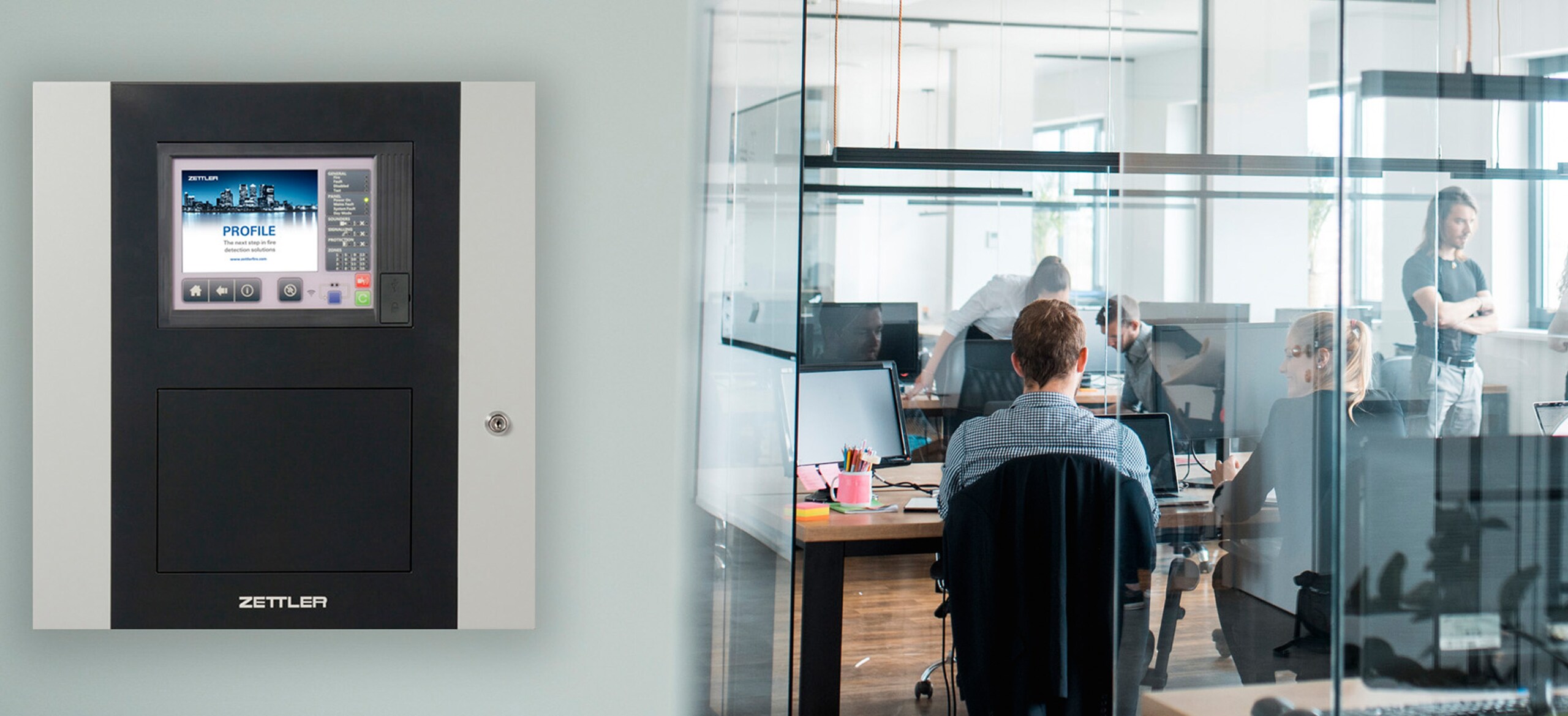 A fire panel on a wall next to an office with people working inside