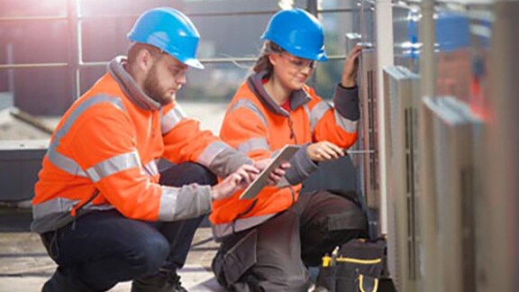 Two mechanics kneeling before a machine as one of them notes down observations