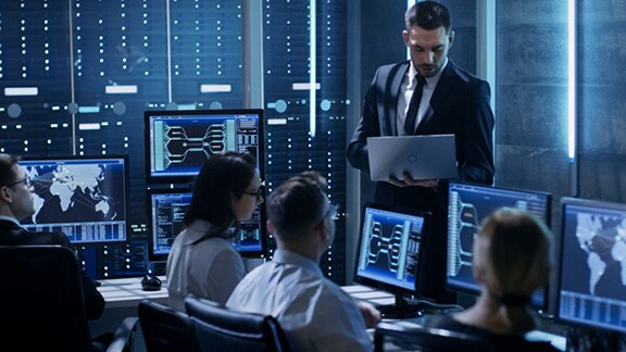 A group of employees seated at computers, while a man stands and holds a laptop