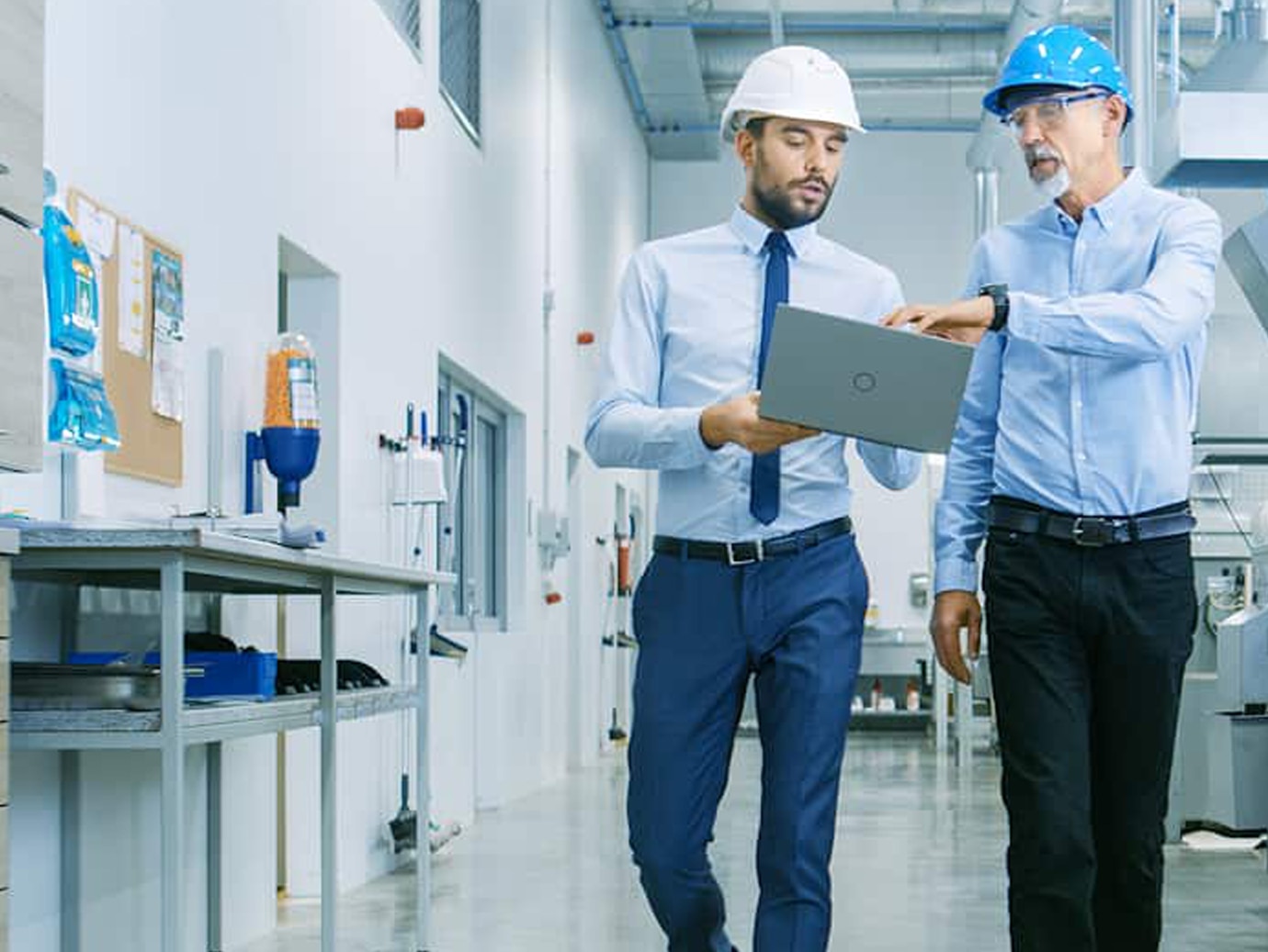 Two male engineers holding a laptop and talking, while walking along a medical facility