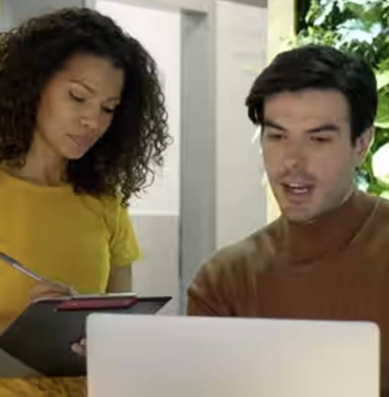 A man looking at a laptop while a woman takes notes beside him